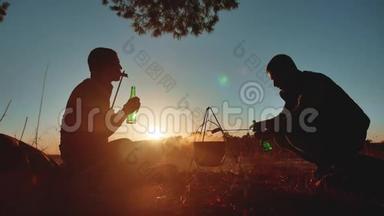 两名游客正坐在篝火晚会的露营生活方式中，在大自然的剪影阳光下日落。 两个男人剪影